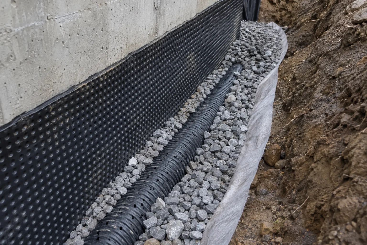 Drain perforé posé dans la pierre nette au pied d’une fondation avec membrane alvéolée